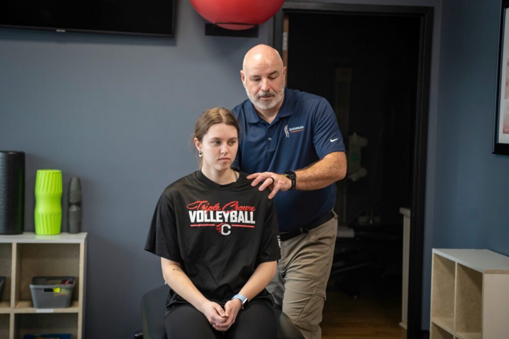 Chiropractor performing shoulder and upper back examination on seated young patient in clinical room